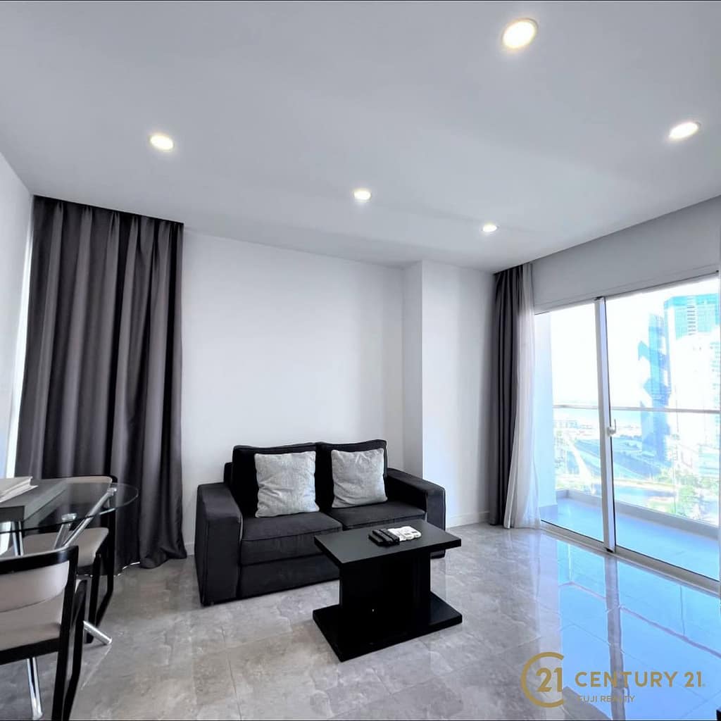 Modern living room at Meridian Koh Pich featuring a black sofa, two cushions, small coffee table, glass dining table with chairs, large window with city view, gray curtains, tiled floor, and recessed ceiling lighting.