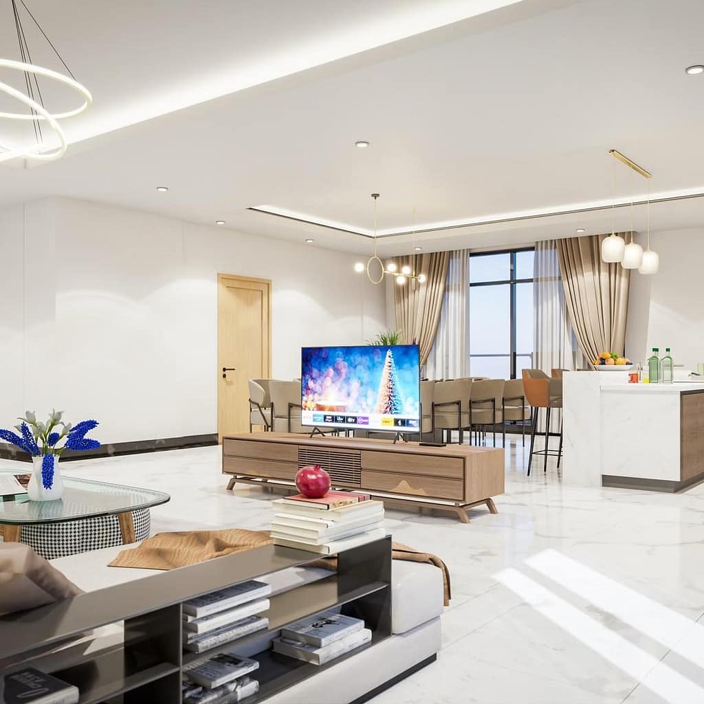 A modern, open-plan living room with a TV on a wooden stand, a marble floor, a cozy sofa, a coffee table with books, and a kitchen area with bar stools and pendant lights in the background.