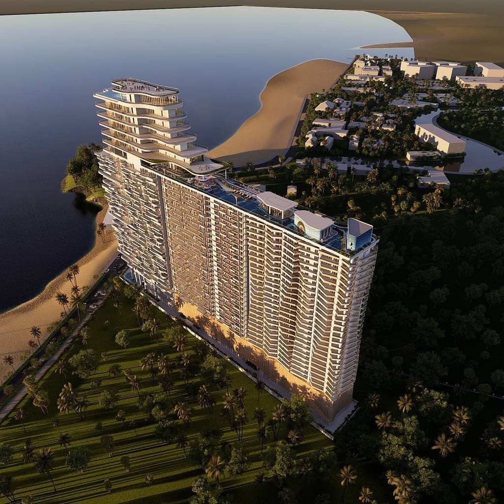 A modern, high-rise building with multiple terraces overlooks a waterfront and sandy beach, surrounded by palm trees and greenery, with smaller buildings in the background.