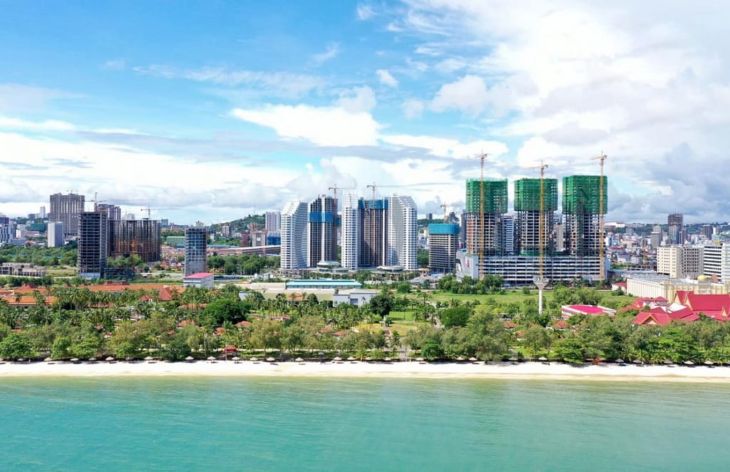 A coastal cityscape in Star Bay Sihanoukville features high-rise buildings under construction, green trees lining the waterfront, and a calm blue sea in the foreground beneath a partly cloudy sky.