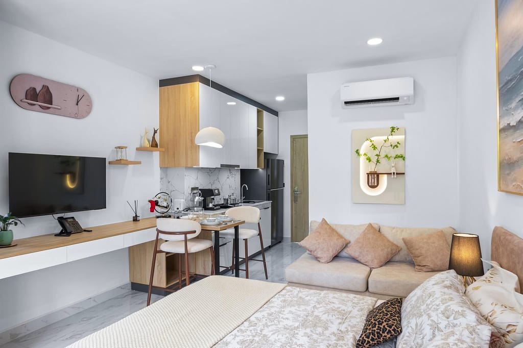 A modern studio apartment with a bed, sofa, wall-mounted TV, wooden desk, small dining area, and a compact kitchen. The space is decorated with plants, neutral tones, and minimalist decor.