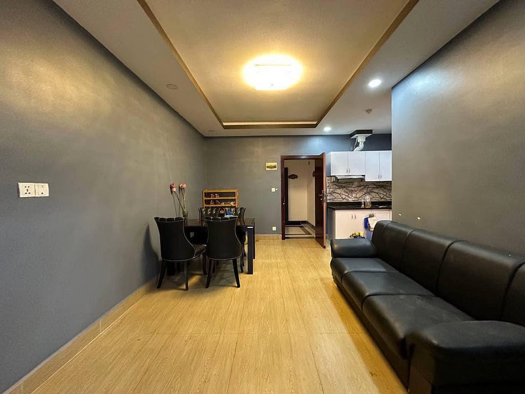A modern condo for sale featuring a living and dining area with gray walls, wood-look flooring, a black sofa, black dining table with chairs, and a view of the compact kitchen. Ceiling lights and minimal decor complete the stylish space.