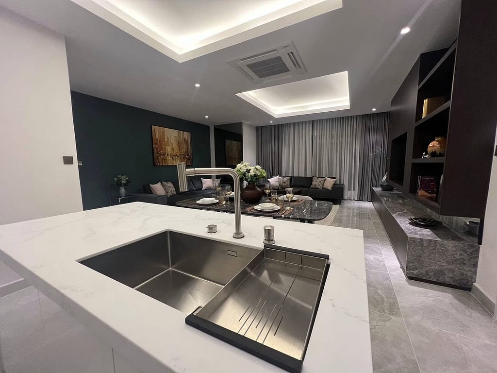 A modern open-concept living room in a stylish 3 bedroom apartment, featuring a marble-topped kitchen island and sink, a cozy seating area with sofa and cushions, coffee table with flowers, and a large TV on a sleek media unit.