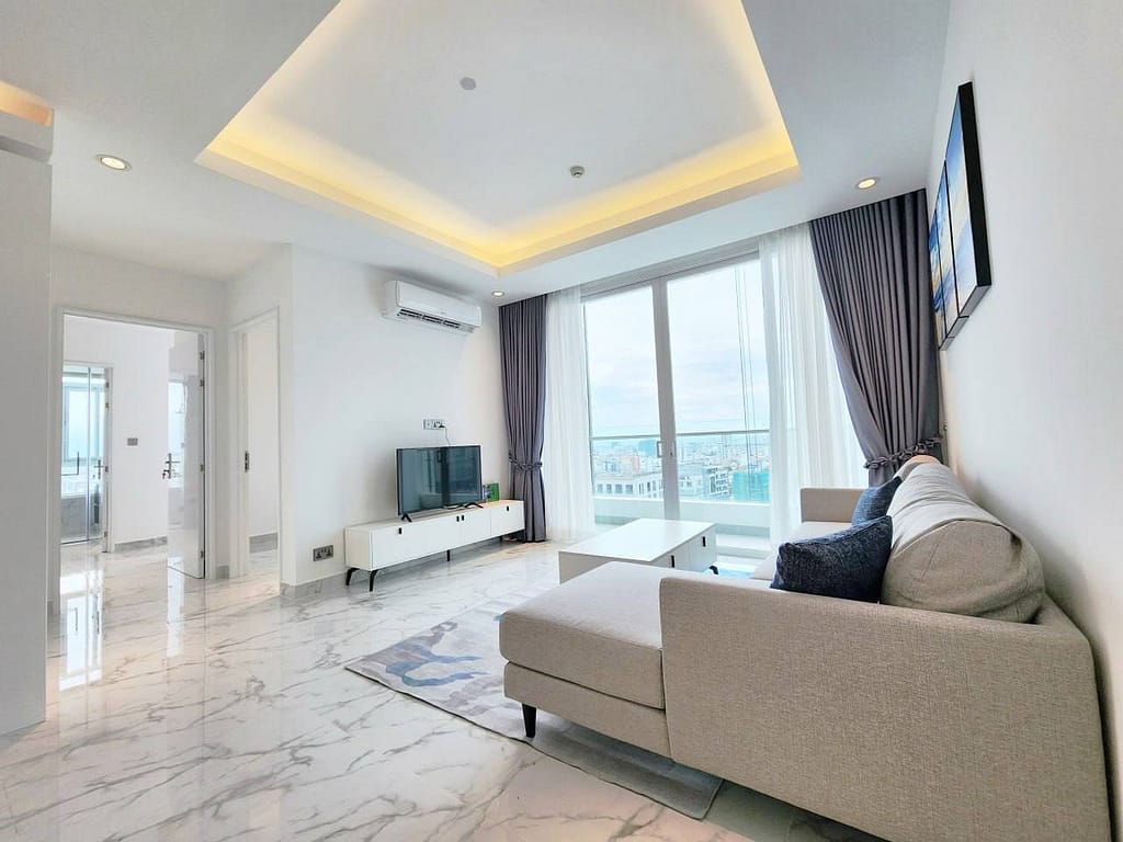 A modern, bright resale apartment living room featuring marble floors, a gray sectional sofa, a white coffee table, a TV on a white stand, large windows with city views, and ceiling lights with recessed lighting.