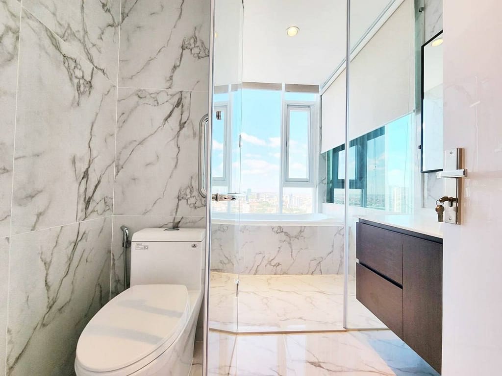Modern bathroom in a resale apartment with marble walls and floors, a toilet on the left, glass shower in the center, and a bathtub by a large window with city views; dark wood vanity on the right. Bright and clean appearance.