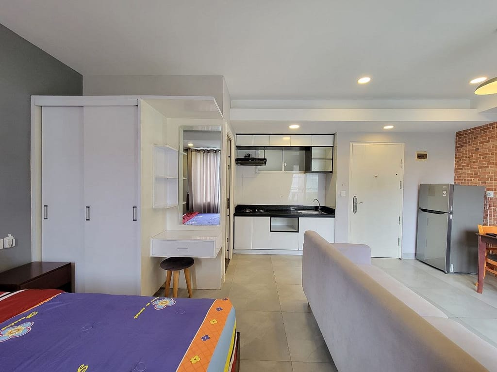 A modern studio apartment with a bed in the foreground, a wardrobe, a small vanity with a stool, a kitchenette, a refrigerator, and a sofa. Natural light enters through a window with curtains.