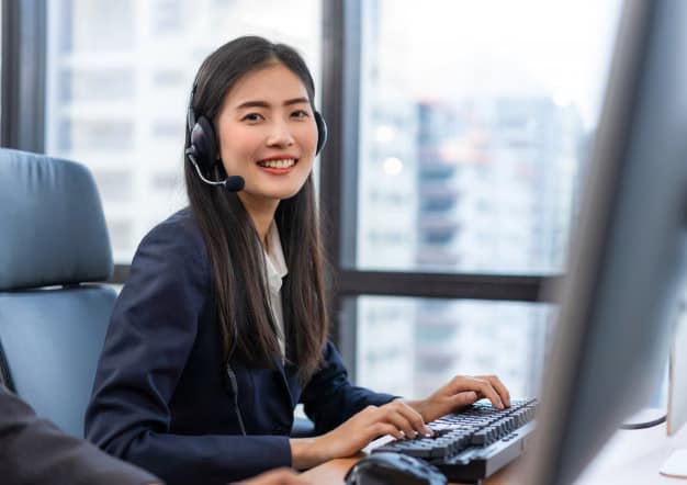 happy_smiling_operator_asian_woman_customer_service_agent_with_headsets A woman wearing a headset and a business suit is smiling at her desk while typing on a keyboard, ready to assist with any contact needs, in a modern office with large windows in the background.